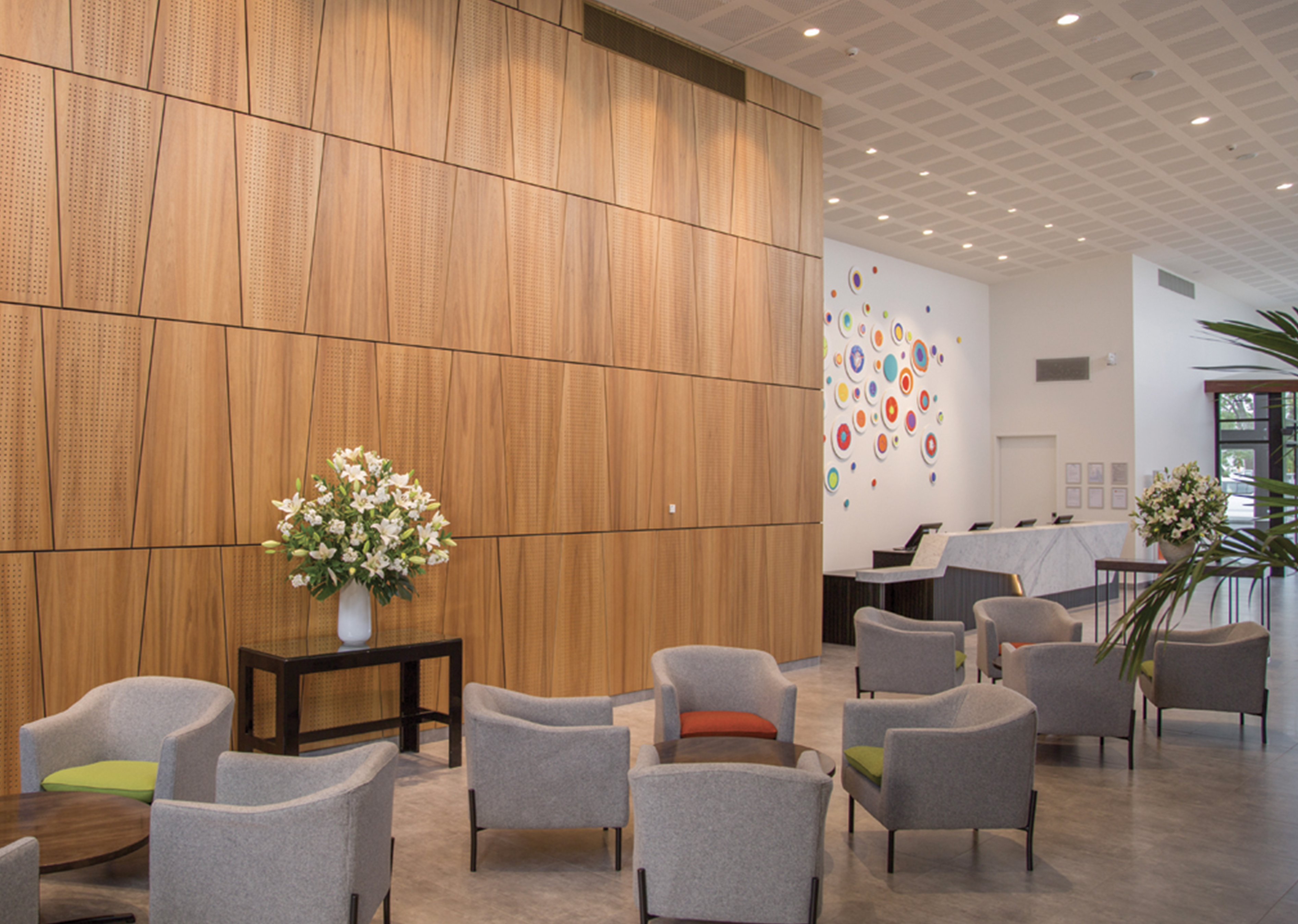 Inside view of hotel lobby interior. Multiple gray chairs and wooden tables for guests.  Ambient lighting with a combination of white paint walls and wood finished walls.