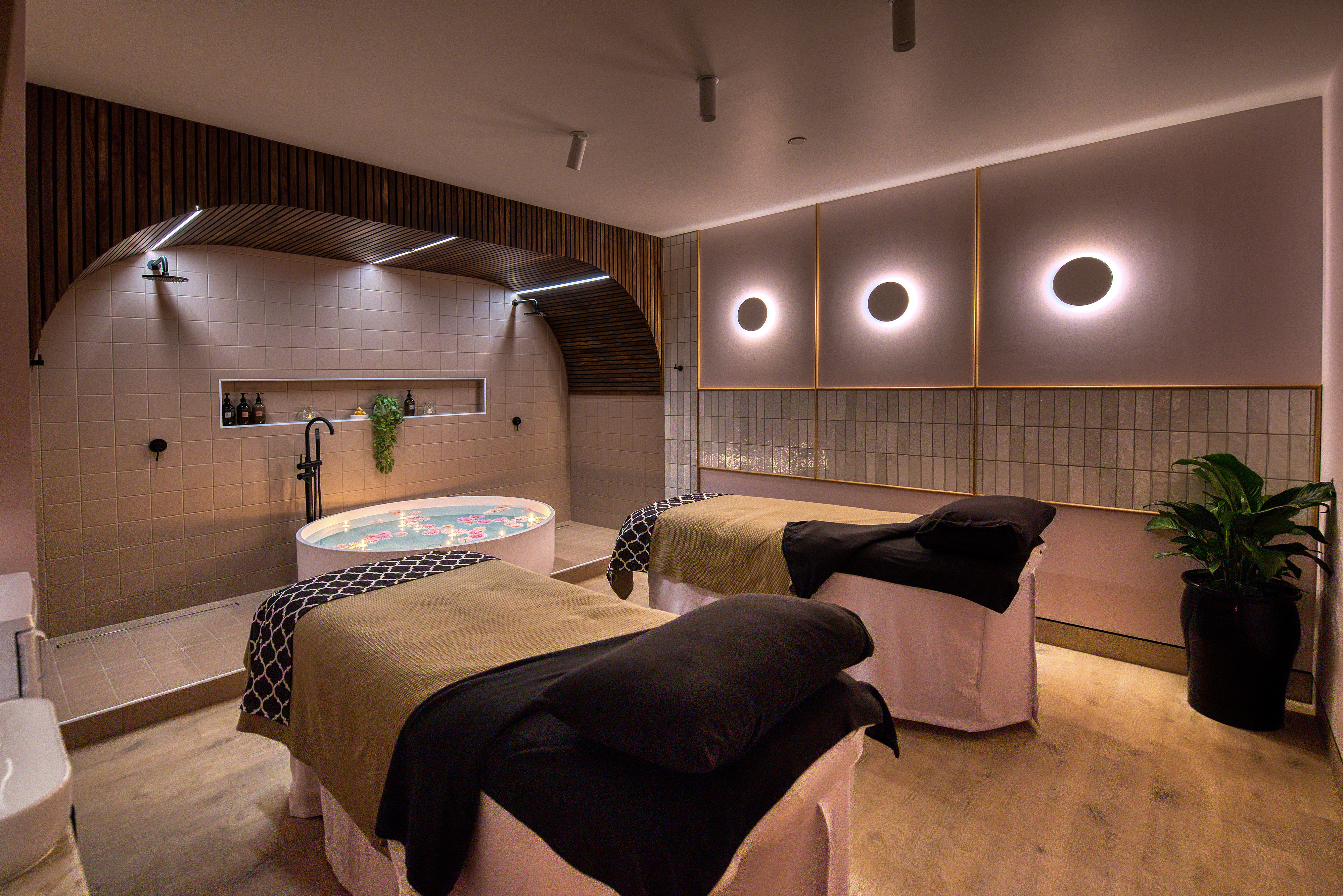 Double treatment spa room with two beds and spa bath in between. Subtle lighting and pink furnishings.