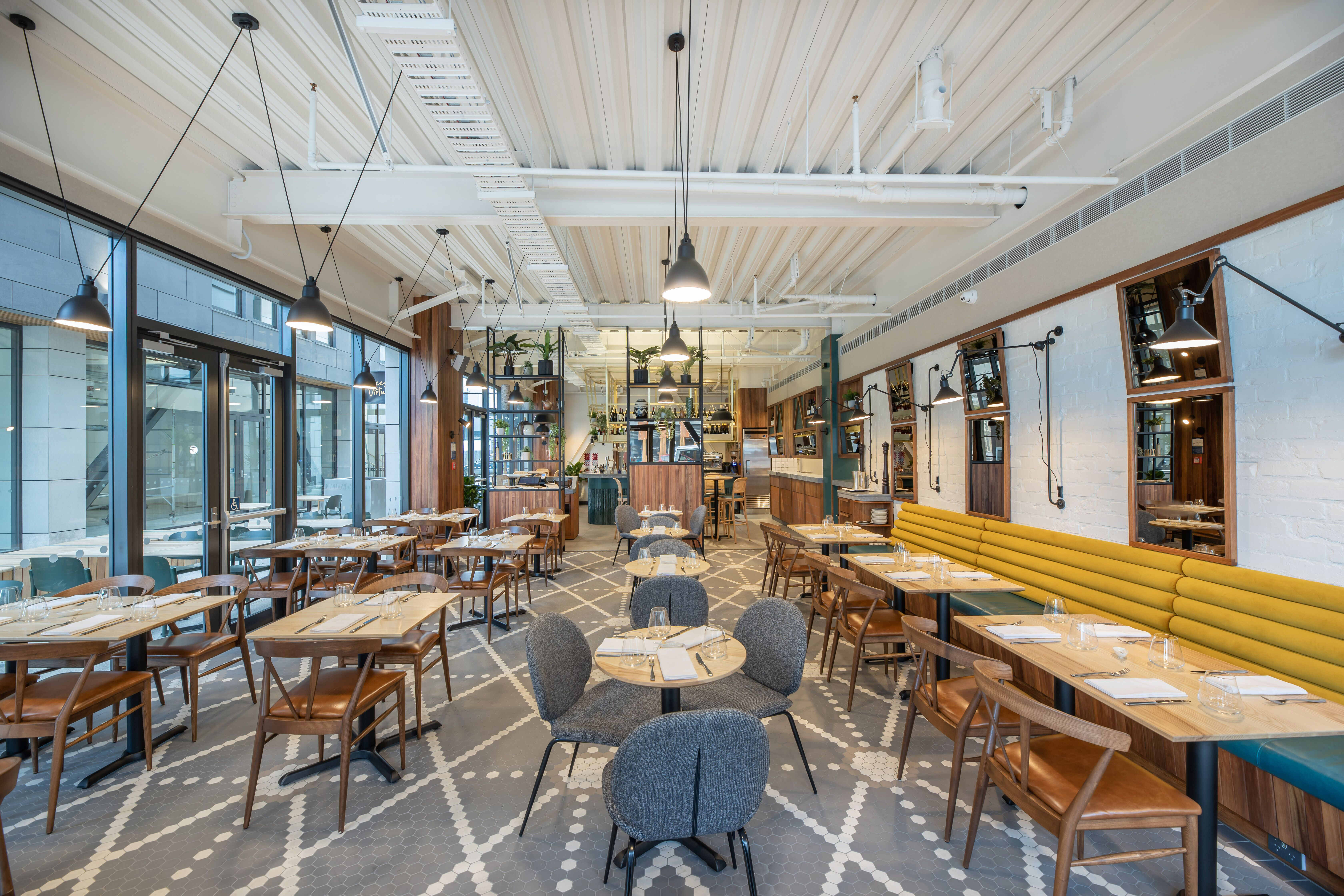 Interior view of a restaurant. Multiple wooden dining tables, couch and chairs, with complete cutlery on every table. light shines through its elegant glass windows. 