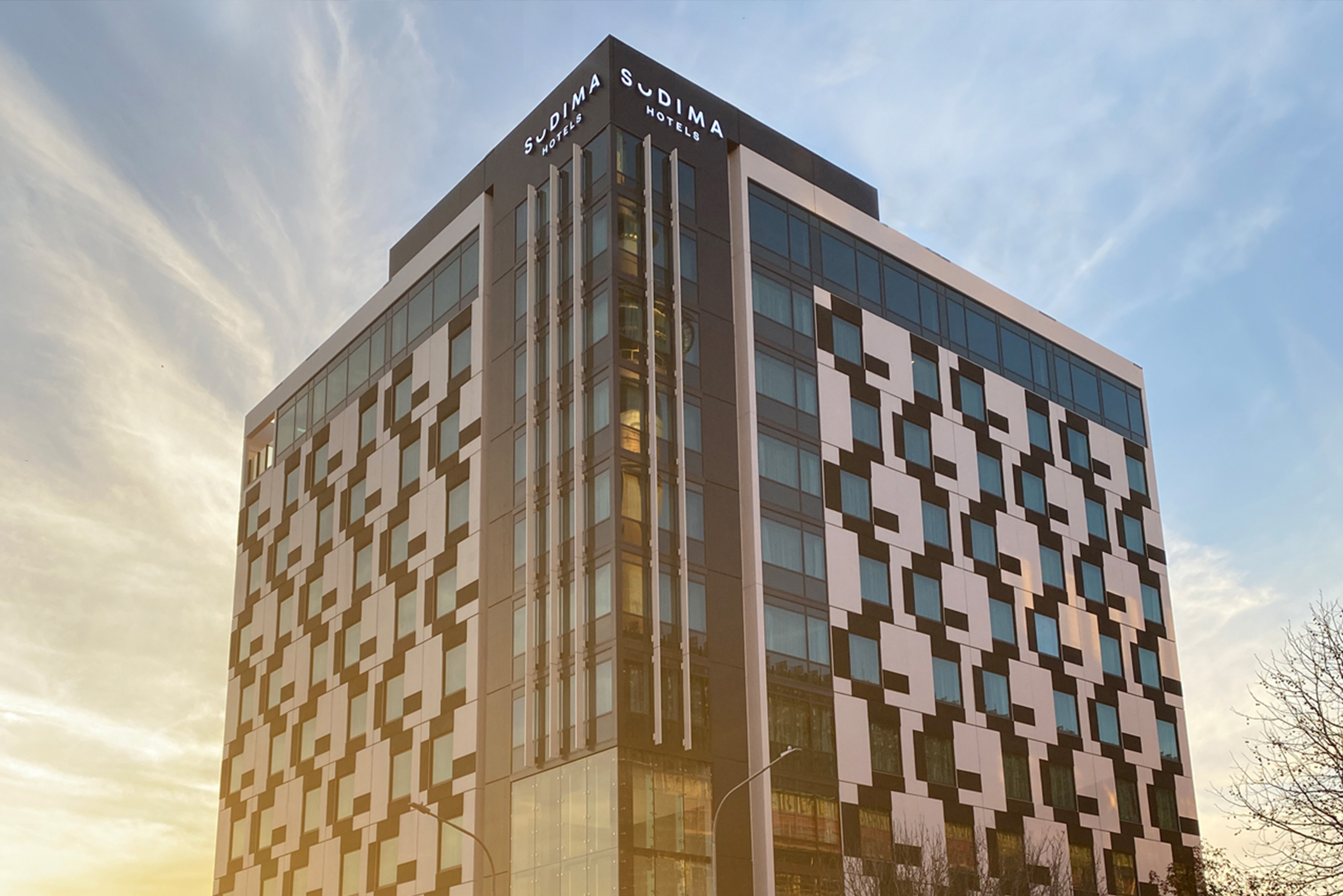 Outside front view of sudina hotel's building. Modern black and cream building to go along with sleek multiple glass panel windows while being illuminated by the sun behind it. 