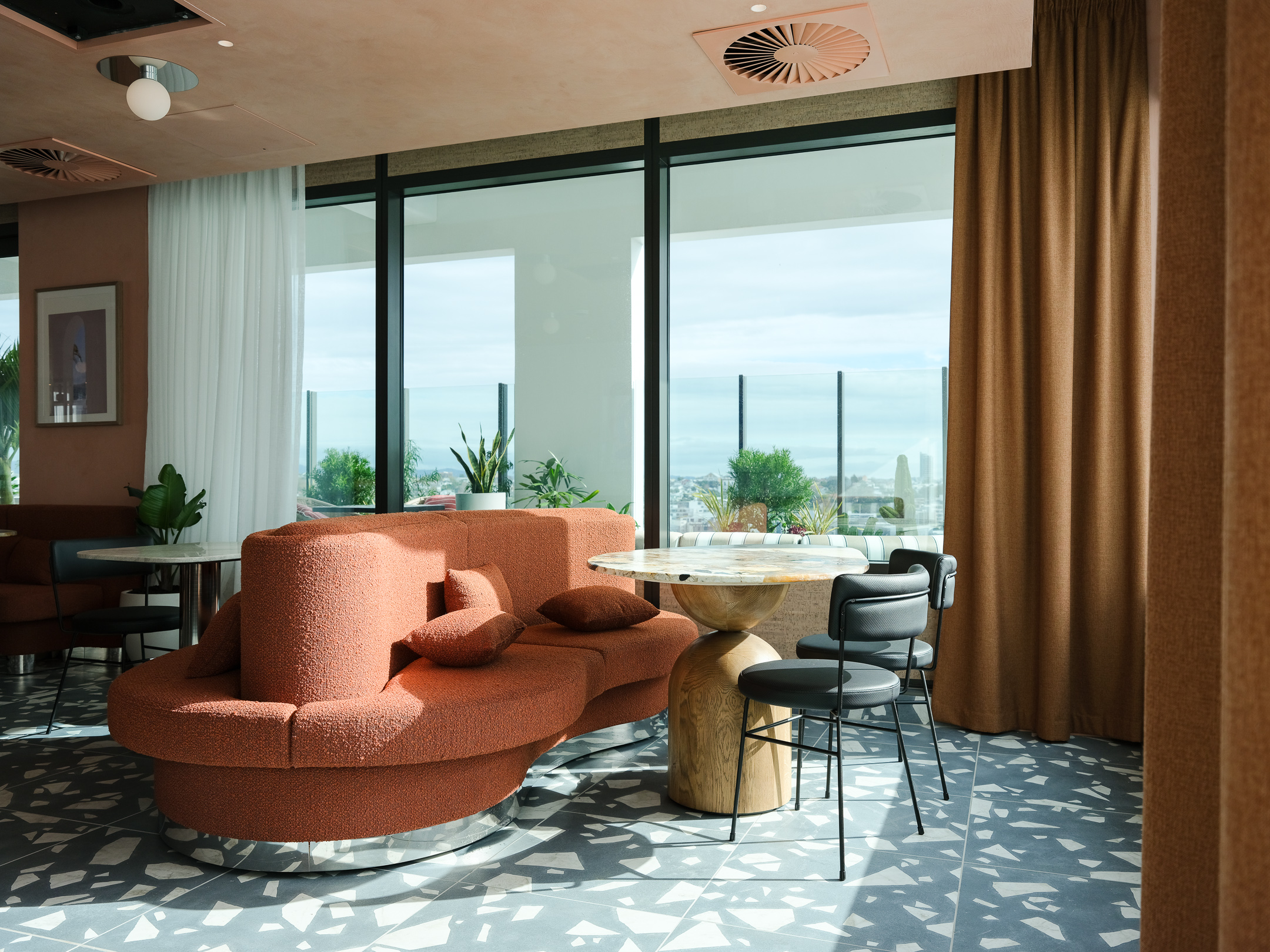 Inside view of a living room interior. A long round orange couch with pillows, black leather chairs, and a round granite table in between. behind it are huge windows with a stunning view outside.